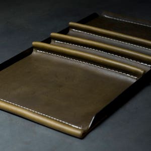 May include: A brown leather tray with five slots for holding items, such as pens or pencils. The tray has a black metal frame and white stitching around the edges.
