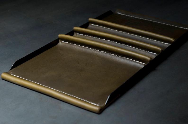 May include: A brown leather tray with five slots for holding items, such as pens or pencils. The tray has a black metal frame and white stitching around the edges.