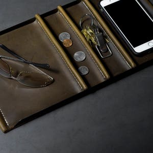 May include: A brown leather tray with multiple compartments. The tray contains a pair of sunglasses, a set of keys, a smartphone, and several coins.