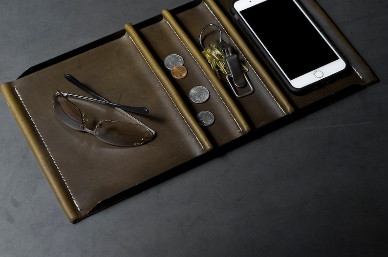 May include: A brown leather tray with multiple compartments. The tray contains a pair of sunglasses, a set of keys, a smartphone, and several coins.