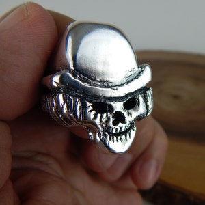 May include: A silver skull ring with a bowler hat on top. The ring is detailed with a textured skull and hat.
