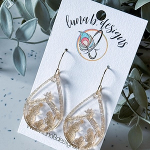 May include: Gold-colored teardrop earrings with a nativity scene design. The earrings are suspended from gold-colored hooks and are displayed on a white card with the text "luna b. designs" and the website address.