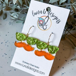 May include: A pair of novelty earrings featuring green glittery sunglasses and orange mustaches. The earrings are attached to gold-colored hoops and displayed on a white card with the text "luna b. designs" and "www.lunaBdesigns.com".