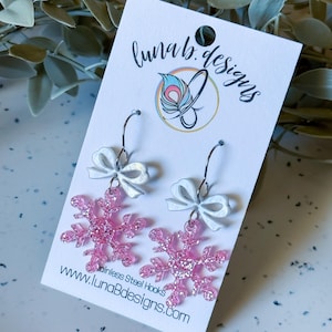 May include: Pink glitter snowflake earrings with white bow accents, hanging from stainless steel hooks. The earrings are displayed on a white card with the text "luna b. designs" and the website address.