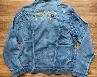 Puede incluir: Chaqueta vaquera azul claro con bordado floral en la espalda. El bordado presenta flores naranjas, amarillas y burdeos con hojas verdes. La chaqueta tiene un cuello clásico y botones.