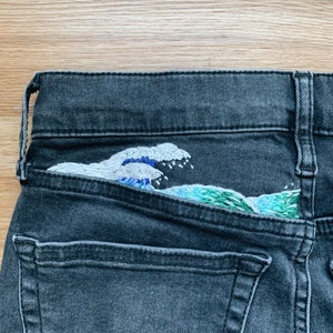 May include: Close-up of a pair of black denim jeans with an embroidered wave design on the back pocket. The wave is depicted in shades of blue, white, and green, adding a pop of color to the dark denim.