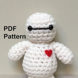 May include: White crocheted amigurumi toy of Baymax from the movie Big Hero 6. The toy has black eyes and a red heart on its chest. The text "PDF Pattern" is visible in the image.