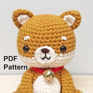 Puede incluir: Juguete de perro amigurumi de crochet en color marrón y blanco. El perro lleva un collar rojo con una campanilla dorada. El juguete está sentado sobre una superficie de madera. Patrón PDF.