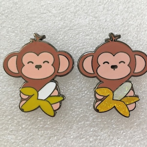 May include: Two brown monkey enamel pins with a yellow banana in their hands. One banana has white glitter and the other has gold glitter.