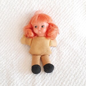 May include: A small, vintage doll with orange hair, brown eyes, and a tan shirt. The doll is wearing black shoes and is lying on a white, textured surface.