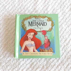 May include: A green book featuring a picture of Ariel from The Little Mermaid on stage. The book is titled "Disney's The Little Mermaid On Stage" and has the text "A Tiny Changing Pictures Book" at the bottom.
