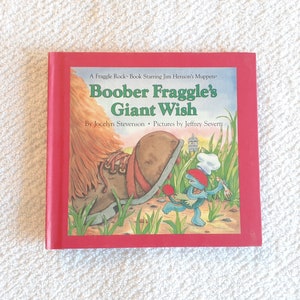 Op de afbeelding: Een rood hardback boek getiteld "Boober Fraggle's Giant Wish" met een Fraggle-personage op de omslag. Het boek maakt deel uit van de Fraggle Rock-serie met Jim Hensons Muppets.
