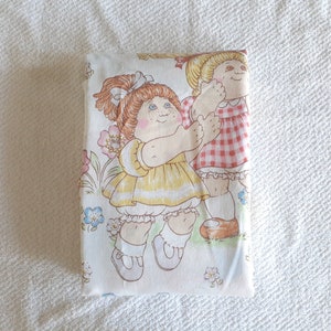 May include: A white fabric with a repeating pattern of two Cabbage Patch Kids dolls. The dolls are wearing dresses and are holding hands. The background is a light pink with flowers.