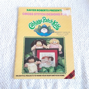 May include: A cross stitch design booklet featuring Cabbage Patch Kids. The cover features a colorful illustration of a Cabbage Patch Kid doll and the text "Cabbage Patch Kids". The booklet includes instructions for creating cross stitch designs of Cabbage Patch Kids.