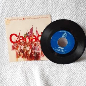 May include: A black vinyl record with a blue label that says "Canada - A Centennial Song - Une Chanson du Centenaire" and features a red maple leaf. The record is on a white surface with a white and red paper sleeve that says "Canada - A Centennial Song - Une Chanson du Centenaire" and features a crowd of people.