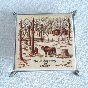 Puede incluir: Un posavasos cuadrado de cerámica con una ilustración en marrón y blanco de una escena de invierno con una cabaña, árboles y dos caballos tirando de un trineo. El texto "Maple Sugaring in CANADA" está impreso en el posavasos.