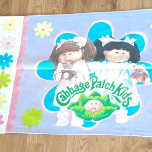 Pode incluir: Uma fronha roxa com uma borda branca e um logotipo Cabbage Patch Kids. O logotipo apresenta duas bonecas com cabelo loiro e castanho, segurando ursinhos de pelúcia. O texto "Cabbage Patch Kids" está escrito em uma fonte verde cursiva.