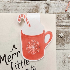 May include: A festive bookmark featuring a red mug design with a snowflake and whipped cream, topped with a candy cane. The text "A Merr Little Meet Cute" is visible. Perfect for book lovers and holiday gifts.