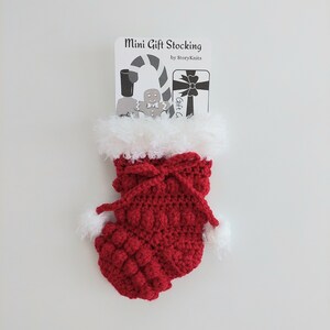 May include: A red crocheted mini gift stocking with white faux fur trim and pom-poms. The stocking has a textured pattern and a bow closure. A tag above the stocking reads "Mini Gift Stocking by StoryKnits" with illustrations of a candy cane, gingerbread man, and gift.