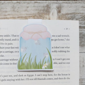 Op de afbeelding: Een grillige boekenlegger met een potontwerp met een lichtblauwe body, een roze deksel en een strik. Binnenin de pot zijn vlinders en gras afgebeeld. Een witte lip steekt uit om het gemakkelijk in een boek te plaatsen.