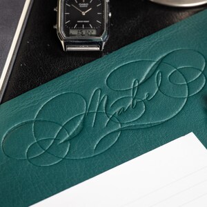 Op de afbeelding: Een teal lederen bureauonderlegger met het woord "Mabe" gegraveerd in een cursief handschrift. Een zilveren horloge en een pen zijn ook zichtbaar op het bureau.