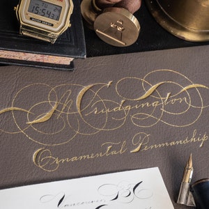 May include: A close-up of a brown leather journal with gold calligraphy that reads "Bridgington Ornamental Penmanship".