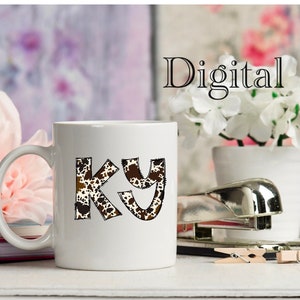 May include: White ceramic mug with a brown and white cow print design that says "KY" on it. The mug is sitting on a table with a pink flower and a stapler.