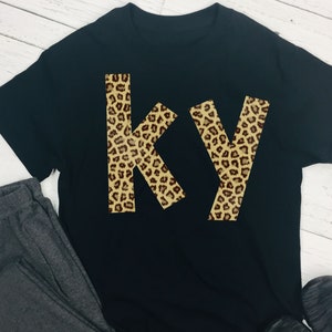 Peut inclure: T-shirt noir avec les lettres "KY" dans un motif léopard.