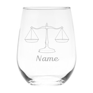 May include: A clear stemless wine glass featuring an etched design of a balanced scale of justice centered on the front. Below the scale, the word Name is etched in a cursive script font. The glass has a rounded body and a smooth rim.