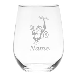 May include: A clear stemless wine glass featuring an etched graphic of a cartoon monkey hanging from a branch while holding a banana. Below the monkey, the word Name is etched in a cursive script font, centered on the front of the glass.