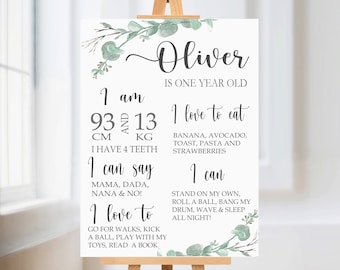 Puede incluir: Un cartel blanco con acentos florales de acuarela verde y el texto "Oliver tiene un año". El cartel enumera la edad, la altura, el peso de Oliver y lo que puede hacer y comer.