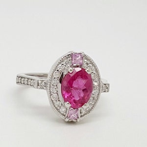 May include: A silver ring with a large oval pink gemstone surrounded by smaller white gemstones. The ring has a delicate band with small white gemstones.