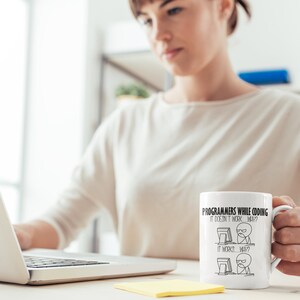 Puede incluir: Taza de cer&aacute;mica blanca con el texto "Programmers While Coding It Doesn't Work... Why? It Works... Why?" e ilustraciones de figuras de palo. La taza la sostiene una persona que trabaja en un port&aacute;til.