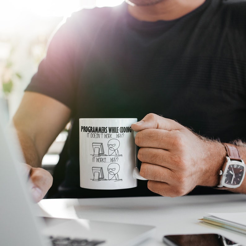Puede incluir: Taza de cer&aacute;mica blanca con el texto "PROGRAMMERS WHILE CODING" y una ilustraci&oacute;n humor&iacute;stica de la experiencia de un programador. La taza tiene un asa y la sostiene una persona.