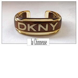 Op de afbeelding: Een bruine en gouden plastic armband met de letters DKNY in reliëf in wit.