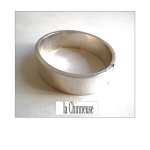 Könnte beinhalten: Ein silberfarbenes, oval geformtes Armreif-Armband. Das Armband hat ein schlichtes, minimalistisches Design mit einer glatten, polierten Oberfläche. Der Text "la Chinneuse" befindet sich in einem weißen Rechteck am unteren Bildrand.