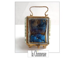 Op de afbeelding: Een kleine, vierkante glazen doos met een gouden frame en een blauwe fluwelen binnenkant. De doos heeft een handvat aan de bovenkant en is versierd met een gouden rand. De doos is gegraveerd met de woorden "Souvenir de Lourdes".
