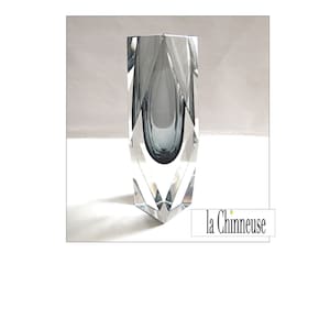 Pode incluir: Um vaso de vidro transparente com um design geométrico e um interior cinza escuro. O vaso está sobre uma superfície branca.