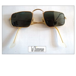 May include: Gold-framed sunglasses with dark lenses. The rectangular frames have a classic design, with a thin gold bridge and temples. The temples have clear, white ear pieces. The text "la Chineuse" is in the lower center.