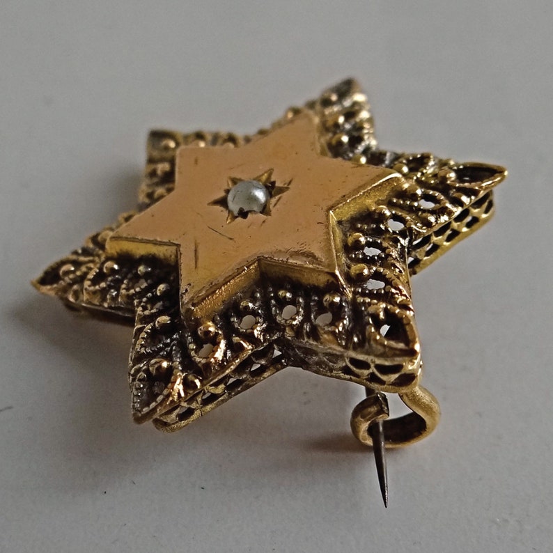 Op de afbeelding: Een gouden Davidster-broche met een parel in het midden. De ster heeft een filigraanontwerp rond de randen.