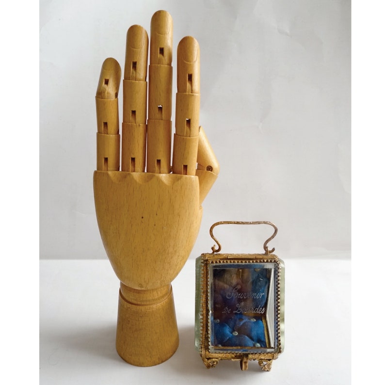 Op de afbeelding: Een houten handmodel met een kleine glazen doos met een gouden frame. De doos heeft een blauwe binnenkant en de tekst "Souvenir de Lourdes" is op de voorkant gedrukt.