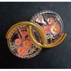 May include: A gold-toned brooch with two clear glass circles featuring pink and white flowers. The flowers are made of delicate, layered paper.