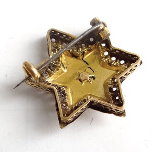 Op de afbeelding: Een gouden stervormige broche met een filigraanontwerp en een kleine, heldere steen in het midden. De broche heeft een speld.