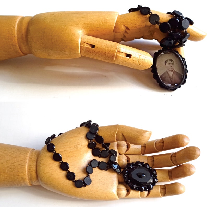 May include: A black jet bead necklace with a cameo pendant featuring a man in a black suit and white shirt. The necklace is displayed on a wooden hand.