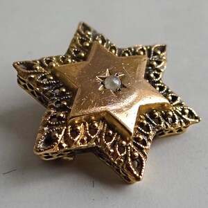 Op de afbeelding: Een gouden Davidster-hanger met een witte parel in het midden. De hanger is versierd met ingewikkelde filigraanwerk.
