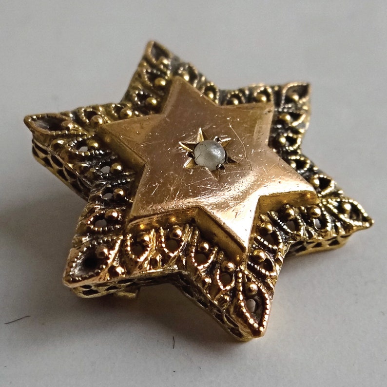 Op de afbeelding: Een gouden Davidster-hanger met een witte parel in het midden. De hanger is versierd met ingewikkelde filigraanwerk.