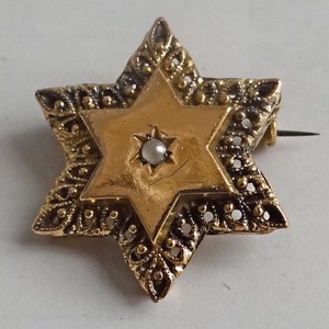 Op de afbeelding: Een gouden Davidster-broche met een parel in het midden. De broche is versierd met ingewikkelde filigraanwerk.