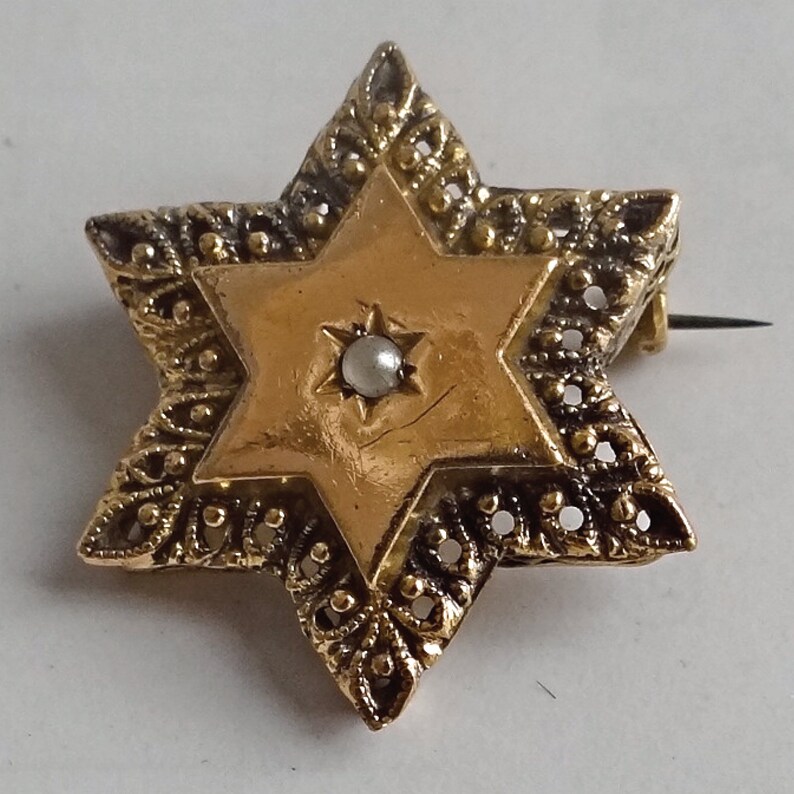 Op de afbeelding: Een gouden Davidster-broche met een parel in het midden. De broche is versierd met ingewikkelde filigraanwerk.