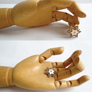 Op de afbeelding: Een houten handmodel dat een gouden stervormige broche vasthoudt met een wit centrum en heldere stenen. De broche is bezet met kleine, heldere stenen rond de randen.