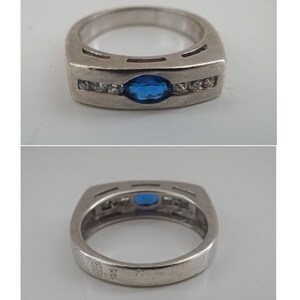 Op de afbeelding: Een zilveren ring met een blauwe ovale edelsteen en een rij heldere edelstenen. De ring is gemarkeerd met "925" aan de binnenkant.